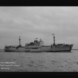 Graues Frachtschiff mit zwei Masten auf ruhiger See; links unten Logo des Internationalen Maritimen Museums.