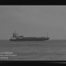 Schwarzweißaufnahme eines Frachtschiffs auf ruhiger See, mit sichtbaren Filmrändern und Museumslogo unten links.