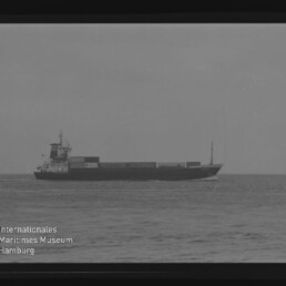Frachtschiff mit aufgestapelten Containern auf ruhiger See, Schwarzweiß-Foto mit Museumslogo unten links.