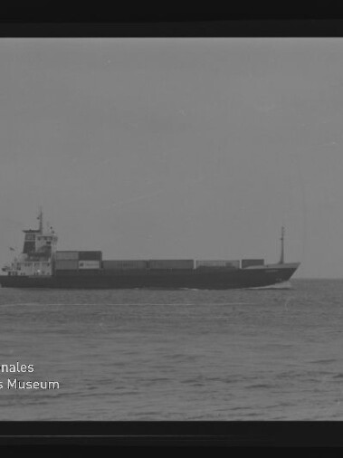 Frachtschiff mit aufgestapelten Containern auf ruhiger See, Schwarzweiß-Foto mit Museumslogo unten links.