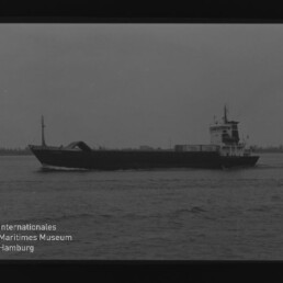 Schwarzweiß-Foto eines Frachtschiffs in Profilfahrt auf ruhiger See, mit Museumslogo und Text unten links.