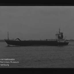Schwarzweiß-Silhouette eines Frachtschiffs mit Ladearm in der Mitte auf ruhiger See, Museumssignet links unten.