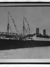 Zweischornsteiniges Passagierschiff mit Wimpelflaggen an Masten am Kai, historische Schwarzweißaufnahme.