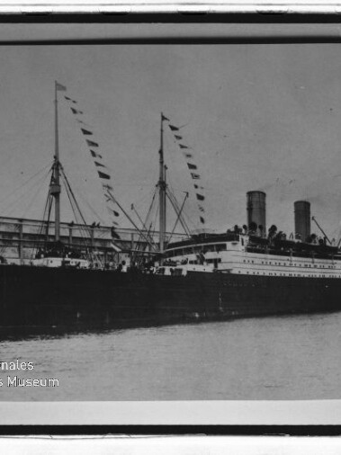Zweischornsteiniges Passagierschiff mit Wimpelflaggen an Masten am Kai, historische Schwarzweißaufnahme.