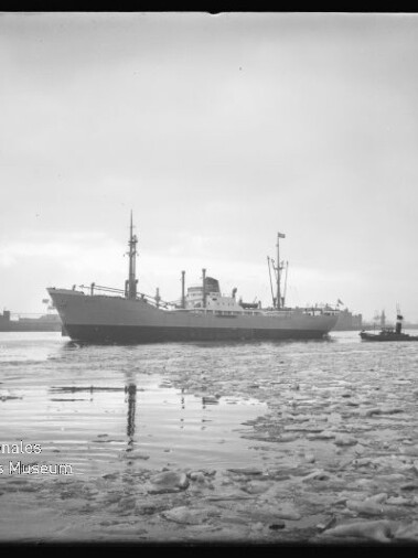Frachtschiff im Hafen, treibende Eisschollen im Vordergrund und ein Schlepper neben dem Schiff.