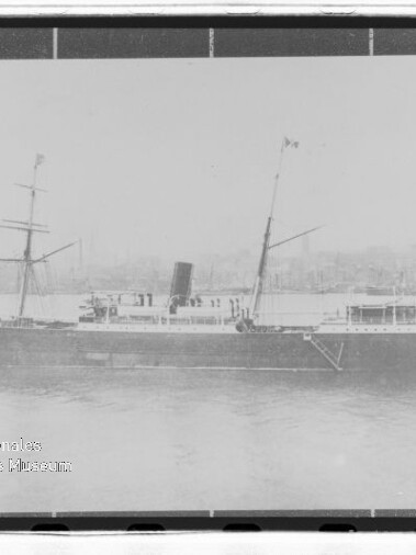 Schwarzweiß-Foto eines historischen Dampfschiffs mit einem Schornstein und zwei Masten, liegend am Hafen vor einer Stadtkulisse.