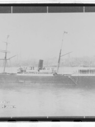 Seitliche Aufnahme eines historischen Dampfschiffs mit zentralem Schornstein und zwei Masten vor einem Hafenhintergrund.