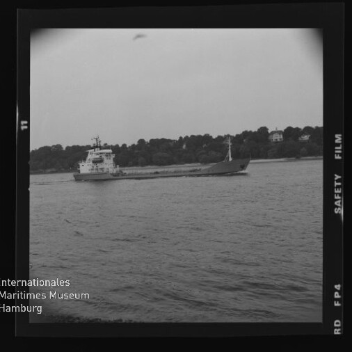 Schwarzweiß-Aufnahme eines Frachtschiffs auf ruhiger See vor bewaldetem Ufer, mit Filmrand und Museumswasserzeichen.