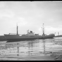 Graustufiges Foto eines Frachtschiffs im Hafen, ruhige, teilweise vereiste Wasserfläche und Hafenanlagen im Hintergrund.