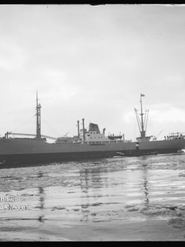 Graustufiges Foto eines Frachtschiffs im Hafen, ruhige, teilweise vereiste Wasserfläche und Hafenanlagen im Hintergrund.