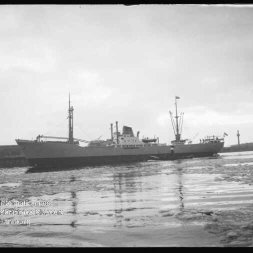 Graustufiges Foto eines Frachtschiffs im Hafen, ruhige, teilweise vereiste Wasserfläche und Hafenanlagen im Hintergrund.