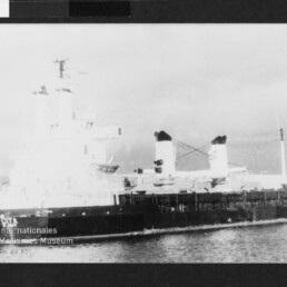 Schwarzweißaufnahme eines großen Frachtschiffs im Hafen mit zwei markanten Schornsteinen und aufragenden Aufbauten.