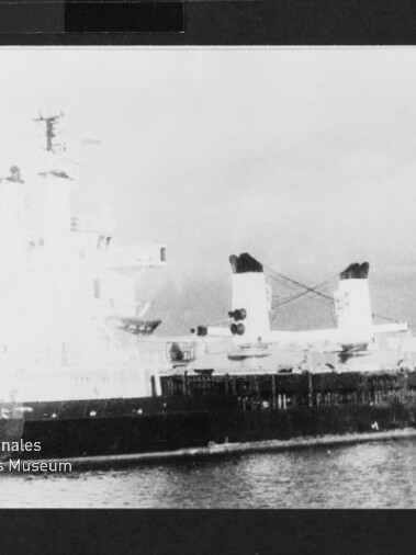 Schwarzweißaufnahme eines großen Frachtschiffs im Hafen mit zwei markanten Schornsteinen und aufragenden Aufbauten.
