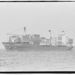 Containerschiff City of Amsterdam mit aufgestapelten Ladungskontainern und zwei Ladekränen auf ruhiger See.