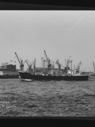 Schwarzweiß-Foto eines Frachtschiffs vor einem Hafen mit zahlreichen Kränen, mit Wasserzeichen Internationales Maritimes Museum Hamburg.