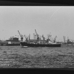 Schwarzweiß-Foto eines Frachtschiffs vor einem Hafen mit zahlreichen Kränen, mit Wasserzeichen Internationales Maritimes Museum Hamburg.
