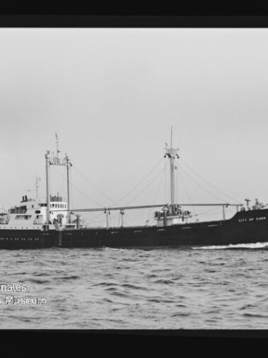 Schwarzweiß-Foto eines Frachtschiffs mit zwei Masten und Ladebäumen, auf ruhiger See fahrend; Museumslogo unten links.