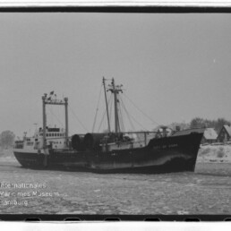 Frachtschiff City of Cork fährt durch vereistes Gewässer nahe Ufer mit Häusern; Schwarzweißaufnahme.