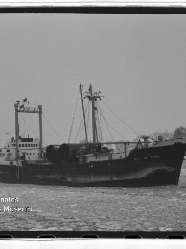 Frachtschiff City of Cork fährt durch vereistes Gewässer nahe Ufer mit Häusern; Schwarzweißaufnahme.