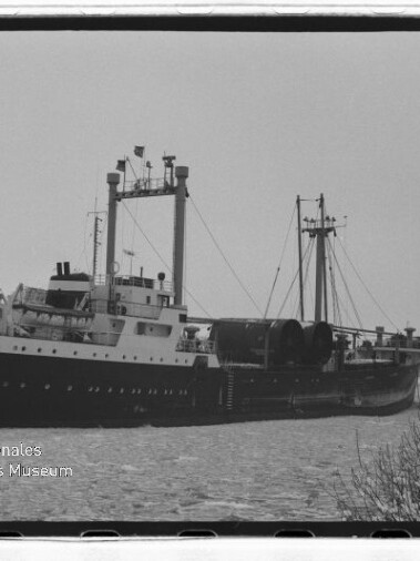 Schwarzweißfoto eines Frachtschiffs mit Masten und großen Rollen an Deck auf ruhigem Wasser; Museumslogo unten links.