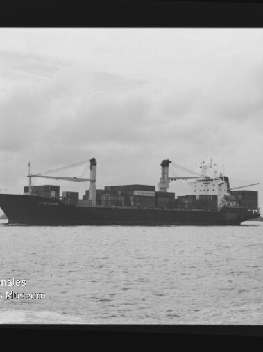 Schwarzweißfoto eines Containerschiffs mit zwei Ladekränen und gestapelten Containern auf ruhiger See unter bewölktem Himmel; Museumslogo links unten.