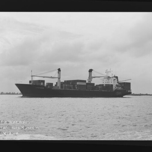 Schwarzweißfoto eines Containerschiffs mit zwei Ladekränen und gestapelten Containern auf ruhiger See unter bewölktem Himmel; Museumslogo links unten.