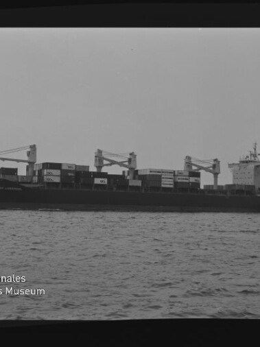 Containerschiff mit geladenen Containerstapeln und Ladekränen auf ruhiger See; Museumslogo unten links.