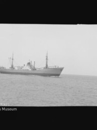 Frachtschiff mit zwei Masten und Aufbauten auf ruhiger See, unten links Logo und Schrift 