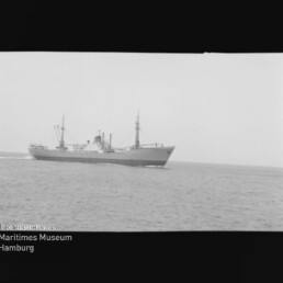 Frachtschiff mit zwei Masten und Aufbauten auf ruhiger See, unten links Logo und Schrift 