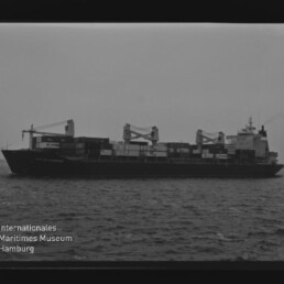 Schwarzweißaufnahme eines Containerschiffs mit Ladekränen und gestapelten Containern auf See, Museumsstempel links unten.