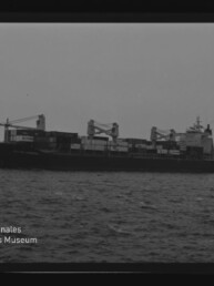 Frachtschiff mit Ladekränen und Containern auf ruhiger See, Schwarzweißaufnahme mit Museumssignet unten links.