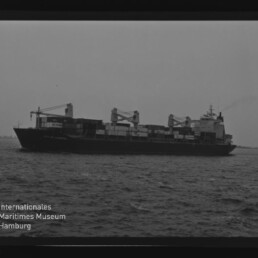 Frachtschiff mit Ladekränen und Containern auf ruhiger See, Schwarzweißaufnahme mit Museumssignet unten links.