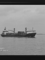 Frachtschiff mit Ladekränen und Containern auf ruhiger See unter bewölktem Himmel; Wasserzeichen Internationales Maritimes Museum Hamburg.