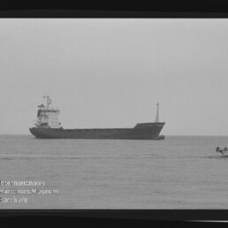 Frachtschiff auf ruhiger See in Schwarzweiß, kleines Beiboot rechts im Bild und Museumslogo unten links.