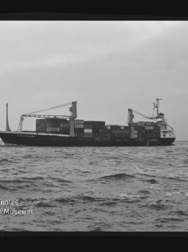 Containerfrachter mit zwei Ladekränen und gestapelten Containern fährt auf welliger See; schwarzweiß-Aufnahme.