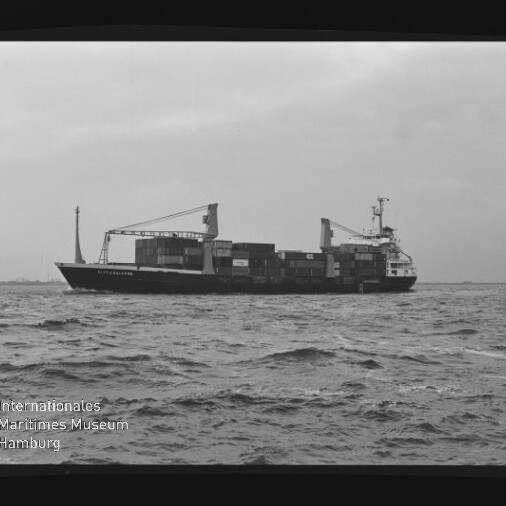 Containerfrachter mit zwei Ladekränen und gestapelten Containern fährt auf welliger See; schwarzweiß-Aufnahme.