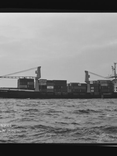 Containerfrachter mit aufgestapelten Ladecontainern und zwei an Deck montierten Kränen, schwarzweiße Aufnahme mit Museumslogo