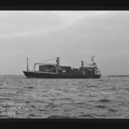 Containerfrachter mit Containern und Ladekränen auf ruhiger See; Museumslogo unten links.