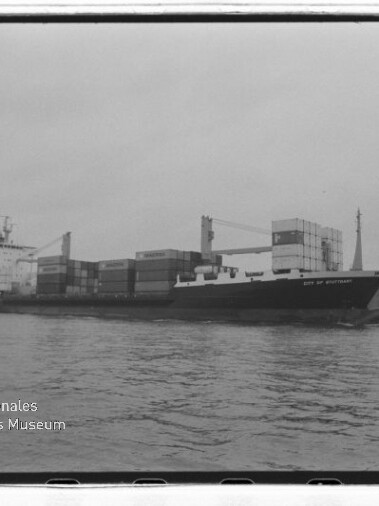 Schwarzweißfoto eines Containerschiffs mit gestapelten Containern und Ladekränen vor einem Ufer, mit Signet Internationales Maritimes Museum Hamburg.