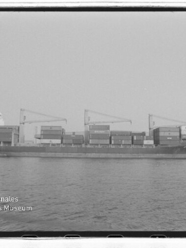 Containerfrachter im Profil mit drei Deckskranen und gestapelten Containern vor Hafen, Wasserzeichen unten links.
