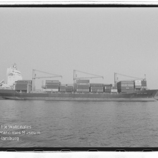 Containerfrachter im Profil mit drei Deckskranen und gestapelten Containern vor Hafen, Wasserzeichen unten links.