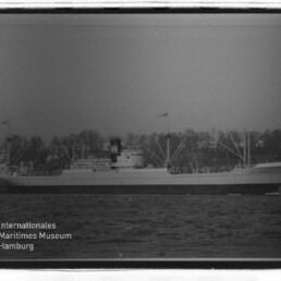 Frachtschiff in Seitenansicht auf See, mit Ladekränen und markantem Schornstein mit den Buchstaben KG