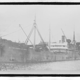 Altes Frachtschiff mit Ladebäumen und Schornstein am Kai; Name am Rumpf teilweise lesbar, Wasserzeichen des Internationalen Maritimen Museums Hamburg