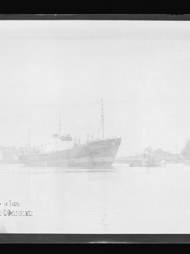 Schwarzweißfoto eines Frachtschiffs im Hafen, Pier links, kleine Boote rechts, Museum-Wasserzeichen unten links.