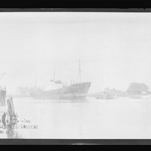 Schwarzweißfoto eines Frachtschiffs im Hafen, Pier links, kleine Boote rechts, Museum-Wasserzeichen unten links.