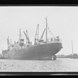 Großes Frachtschiff mit Ladekränen und Signalflaggen im Hafen, Wasser im Vordergrund; Schriftzug Internationales Maritimes Museum Hamburg
