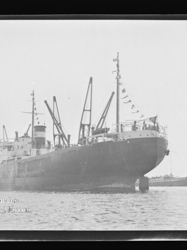 Großes Frachtschiff mit Ladekränen und Signalflaggen im Hafen, Wasser im Vordergrund; Schriftzug Internationales Maritimes Museum Hamburg
