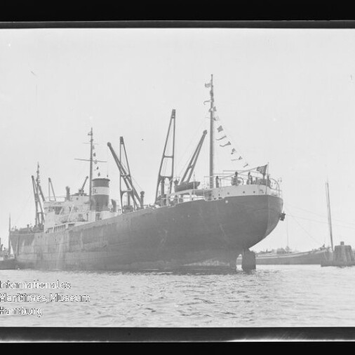 Großes Frachtschiff mit Ladekränen und Signalflaggen im Hafen, Wasser im Vordergrund; Schriftzug Internationales Maritimes Museum Hamburg