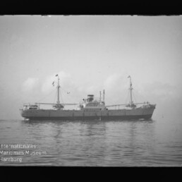Seitenansicht eines Frachtschiffs mit zwei Masten und zentralen Aufbauten auf ruhiger See, schwarzweiß, mit Museumssignet.