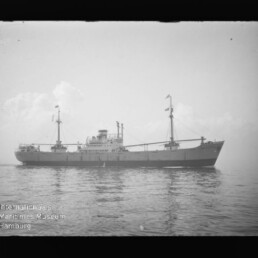 Frachtschiff in Seitenansicht auf ruhiger See, mittige Aufbauten und zwei Masten; schwarzweiß-Foto mit Museumswasserzeichen.
